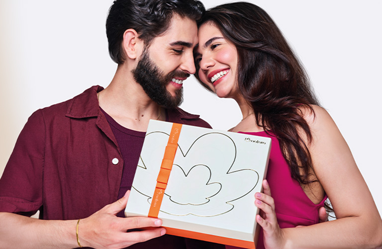 Foto sobre fondo blanco de una pareja abrazada, sosteniendo una caja de regalo blanca para mujer con cinta naranja.